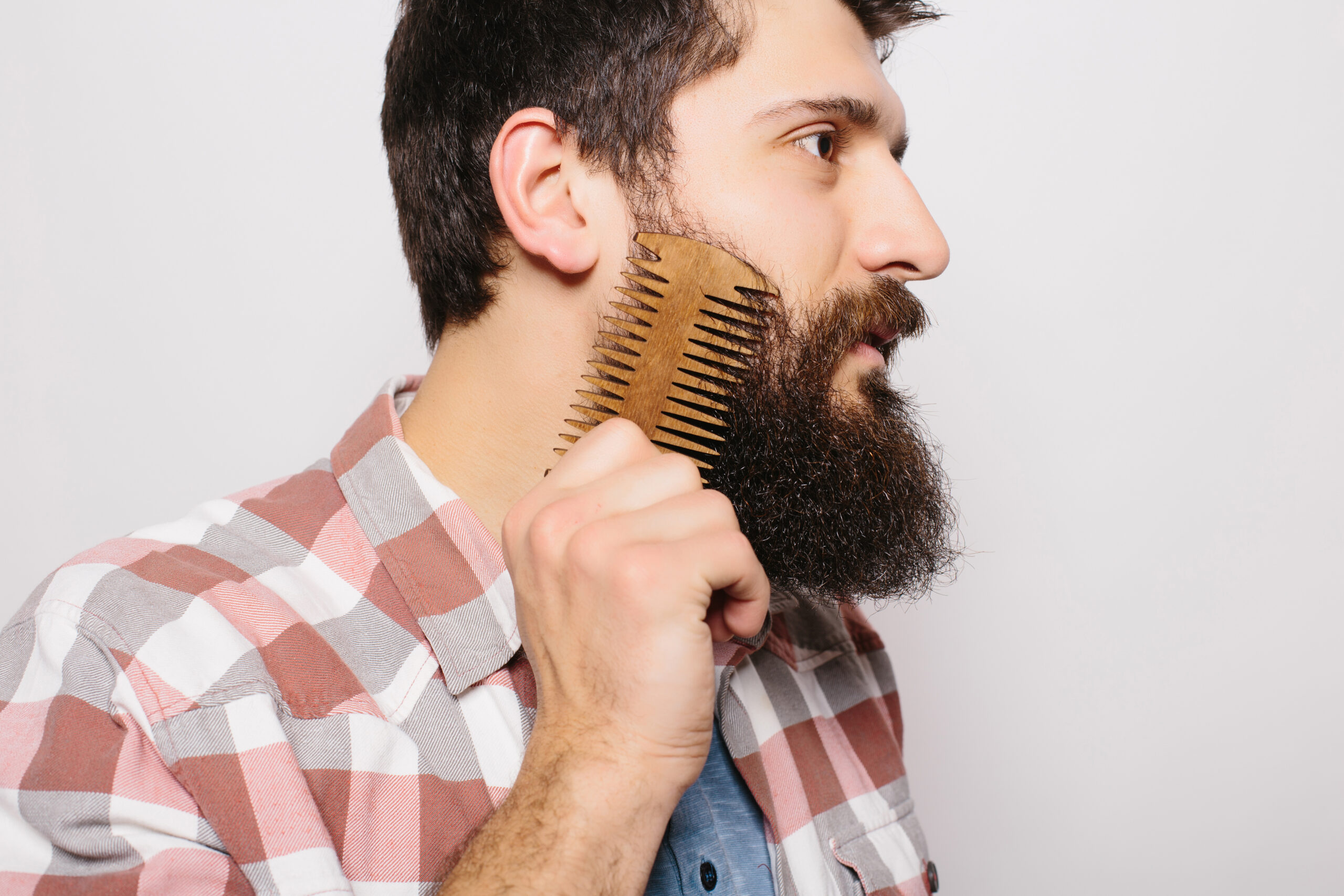 Portrait of young attractive redhead hipster male with serious and confident look, holding wooden comb and doing his thick beard. Stylish bearded barber in checkered shirt combing in salon. Horizontal